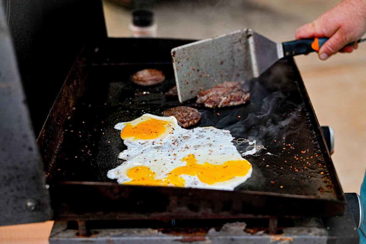Eggs and smash burger patties cooking on a flat top griddle with a wide spatula