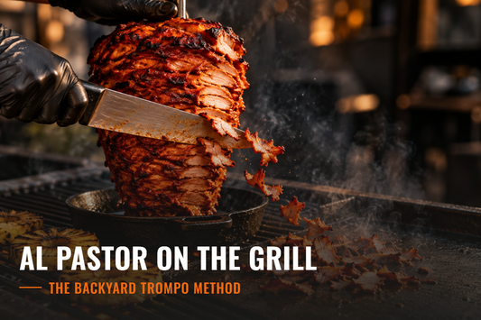 A chef’s knife shaving thin, crispy, red-edged slices of al pastor pork off a vertical trompo stack sitting in a cast iron skillet on a grill grate